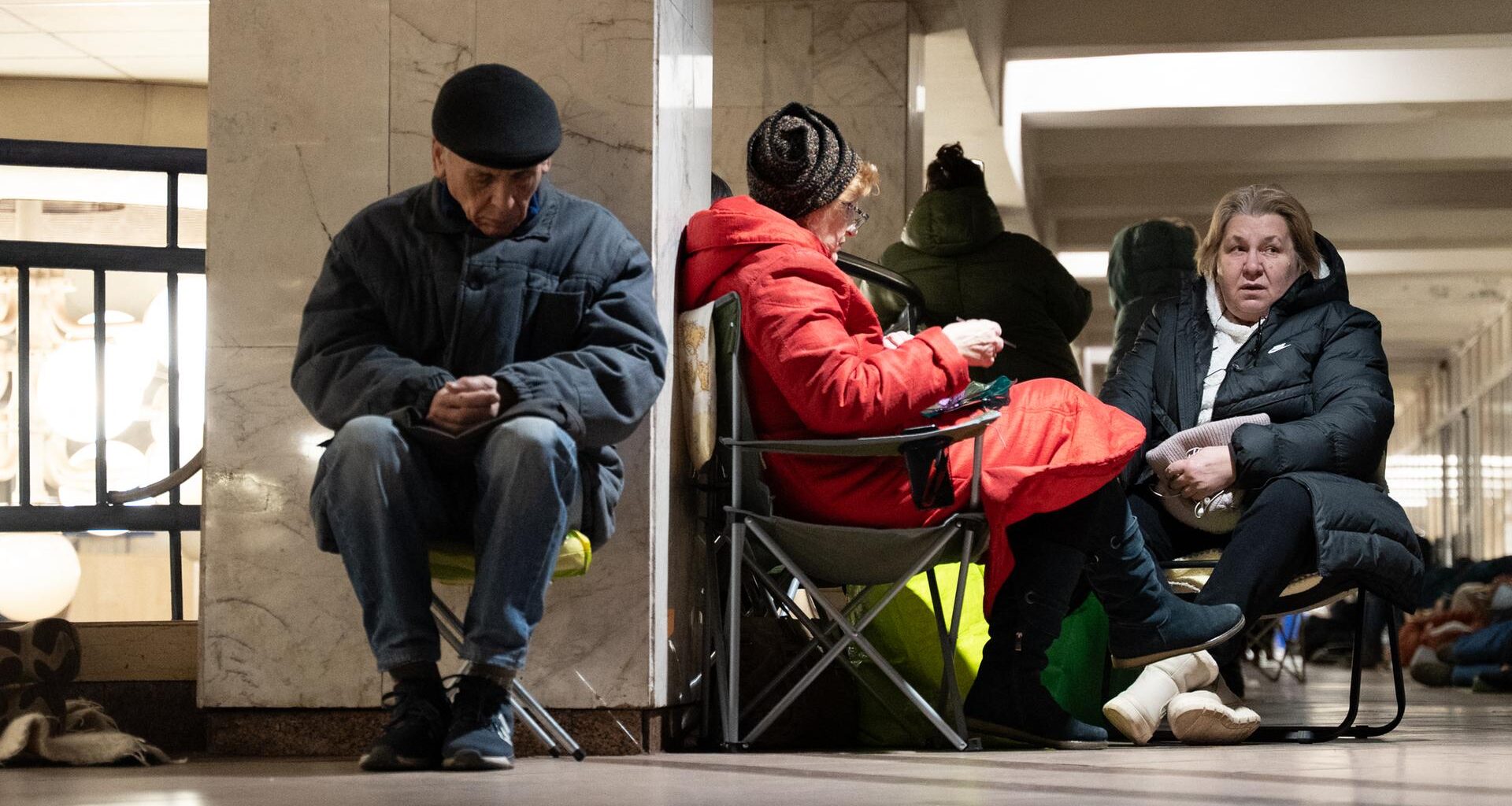 Ein älterer Mann sitzt in Winterkleidung auf einem Klappstuhl an eine Wand gelehnt und scheint zu schlafen. Neben ihm zwei Frauen auf Klappstühlen.