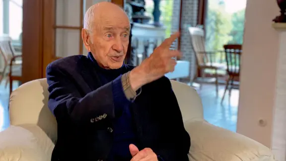 Armin Mueller-Stahl sitzt auf einem Sessel und gestikuliert mit einer Hand, während er spricht.