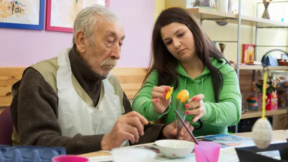 Eine Pflegerin hilft einem älteren Mann beim Bemalen von Ostereiern an einem Tisch