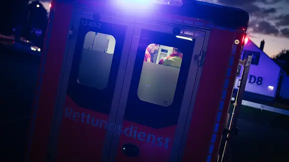 Ein Rettungswagen bei Dunkelheit, durch dessen Heckfenster zwei Rettungssanitäter im Innenraum zu sehen sind.