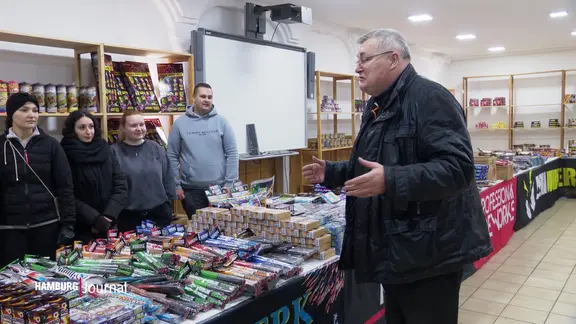 Ladenbesitzer Oliver Graetzer steht vor der Auslage an Feuerwerkskörpern, Böllern und Raketen und spricht mit einer Gruppe junger Personen.