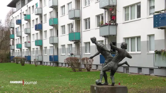 Fassade eines Mehrfamilienhauses mit vielen Balkonen, davor steht eine Statue zweier fußballspielender Personen.