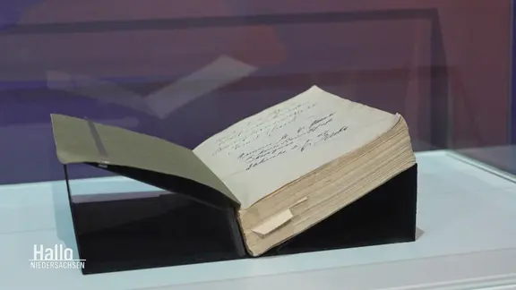 Ein altes aufgeklapptes Buch in einer Glasvitrine. | Screenshot Ein altes aufgeklapptes Buch in einer Glasvitrine.