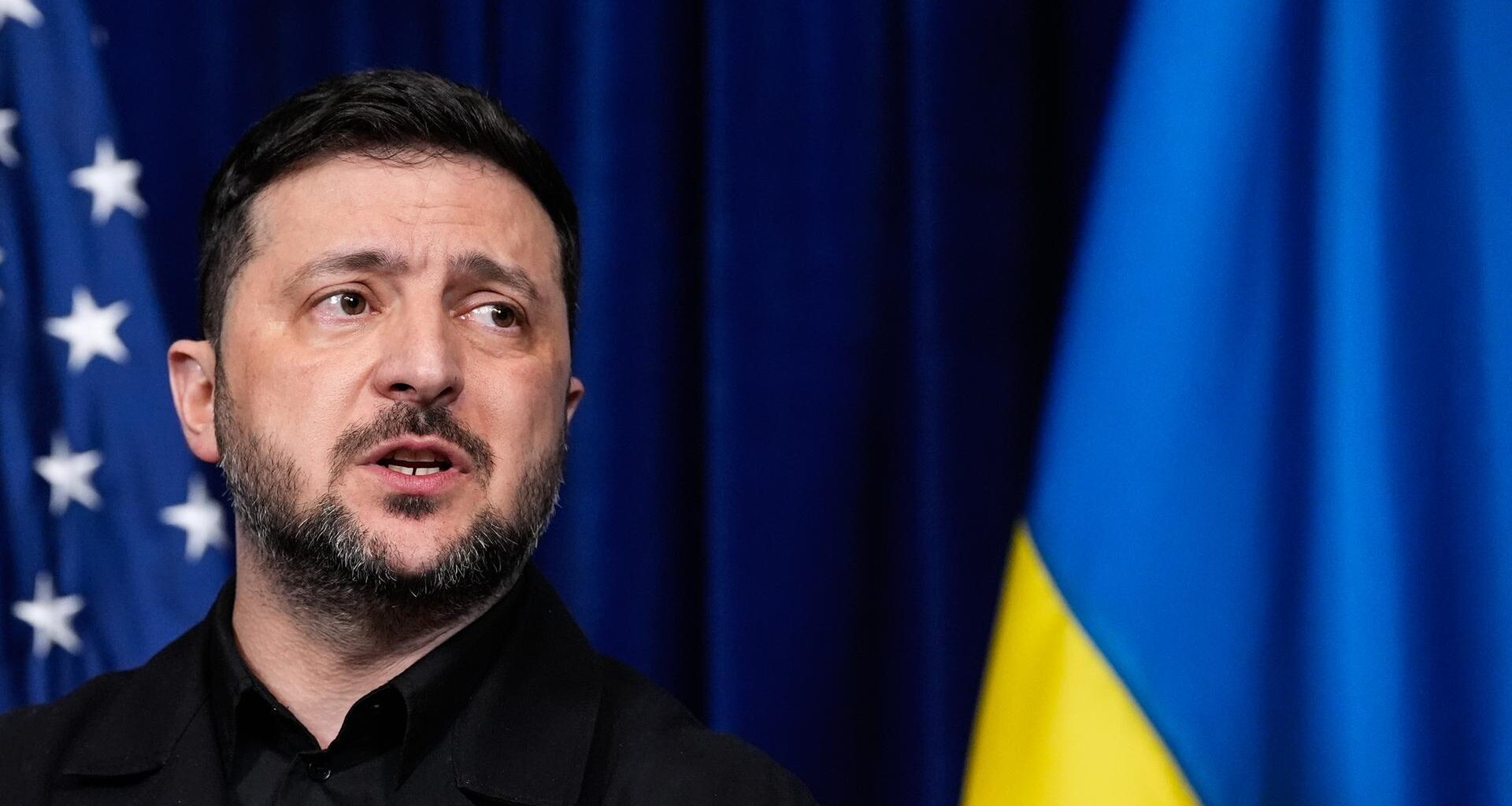 Wolodymyr Selensky spricht während einer Pressekonferenz, dahinter zu sehen sind eine amerikanische und eine ukrainische Flagge.