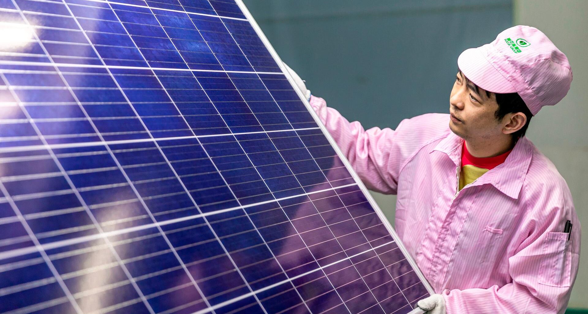 Ein chinesischer Mann hält ein großes Solarmodul in den Händen und schaut prüfend darauf. Er trägt einen rosafarbenen Arbeitsoverall und Mütze.
