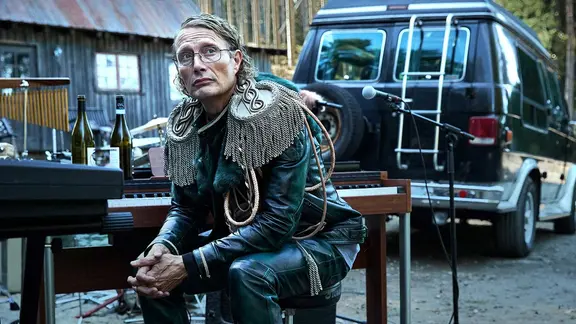 Mads Mikkelsen sitzt mit dem Rücken zu einem Mikrophon und einem Van neben einem Tisch mit Weinflaschen