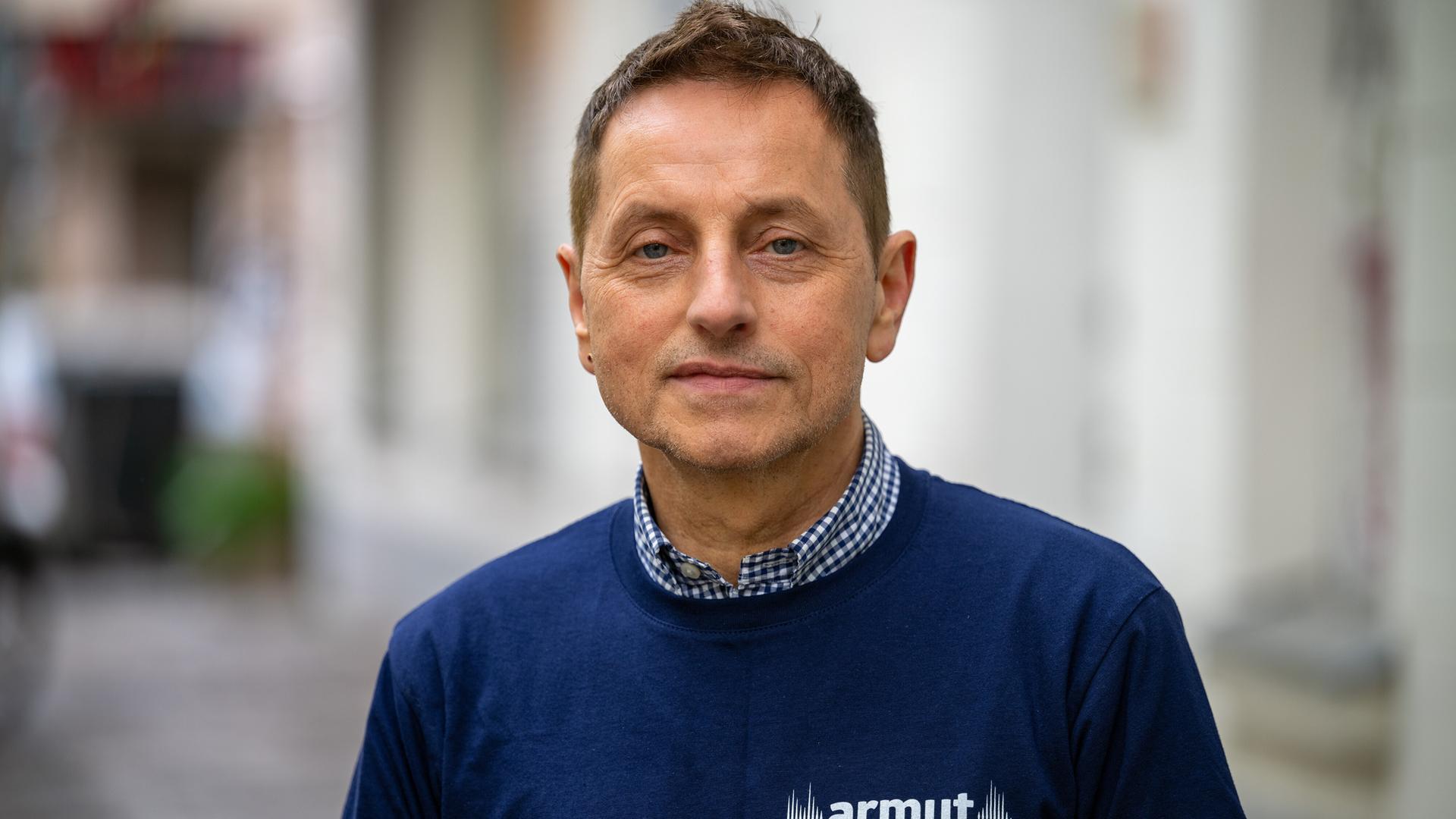 Thomas de Vachroi lächelt in die Kamera. Er trägt ein blaues Shirt mit der Aufschrift "Armut eine Stimme geben".