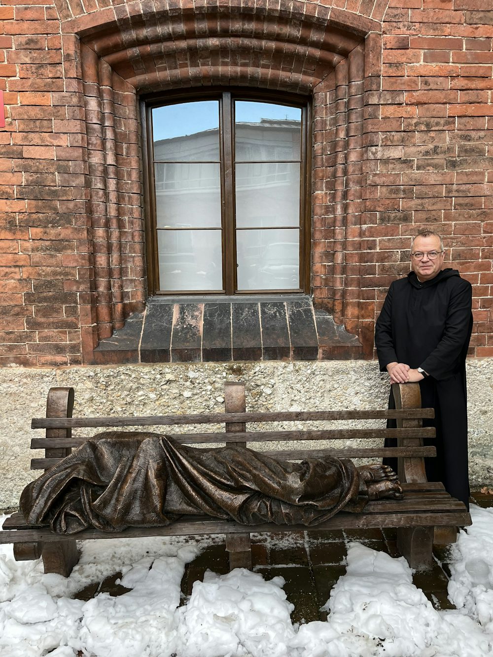 Frater Emmanuel Rotter hat die Obdachlosenhilfe von St. Bonifaz gegründet und leitet sie bis heute. „Homeless Jesus“ heißt das Kunstwerk im tief verschneiten Hof der Benediktinerabtei, bei dem er steht. Die Bronze-Skulptur zeigt den in eine Decke eingeschlagenen Christus und soll an das Jesus-Wort aus dem Matthäus-Evangelium erinnern: „Ich war fremd und obdachlos und ihr habt mich aufgenommen.“