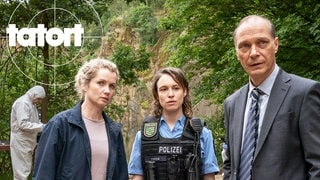 Szenenbild aus dem Tatort „Siebenschläfer“ aus Dresden: Vier Personen stehen an einem Tisch mit Beweismitteln, im Hintergrund arbeiten zwei weitere mit Schutzanzügen. Dichte Bäume umgeben die Szene.