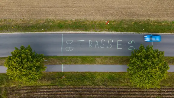 Ein Bild aus der Vogelperspektive: Ein blaues Auto fährt über eine niedersächsische Landstraße. Auf dem Asphalt steht "DB Trasse" geschrieben. | picture alliance/dpa | Philipp Schulze, Philipp Schulze Ein Bild aus der Vogelperspektive: Ein blaues Auto fährt über eine niedersächsische Landstraße. Auf dem Asphalt steht "DB Trasse" geschrieben.