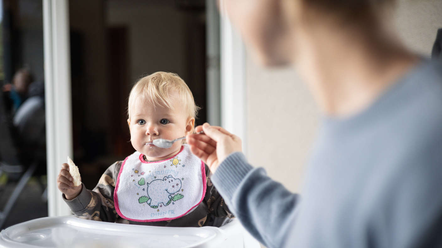 Mother spoon feeding her baby boy child in baby high chair with fruit puree at dinning table at home. Baby solid food in