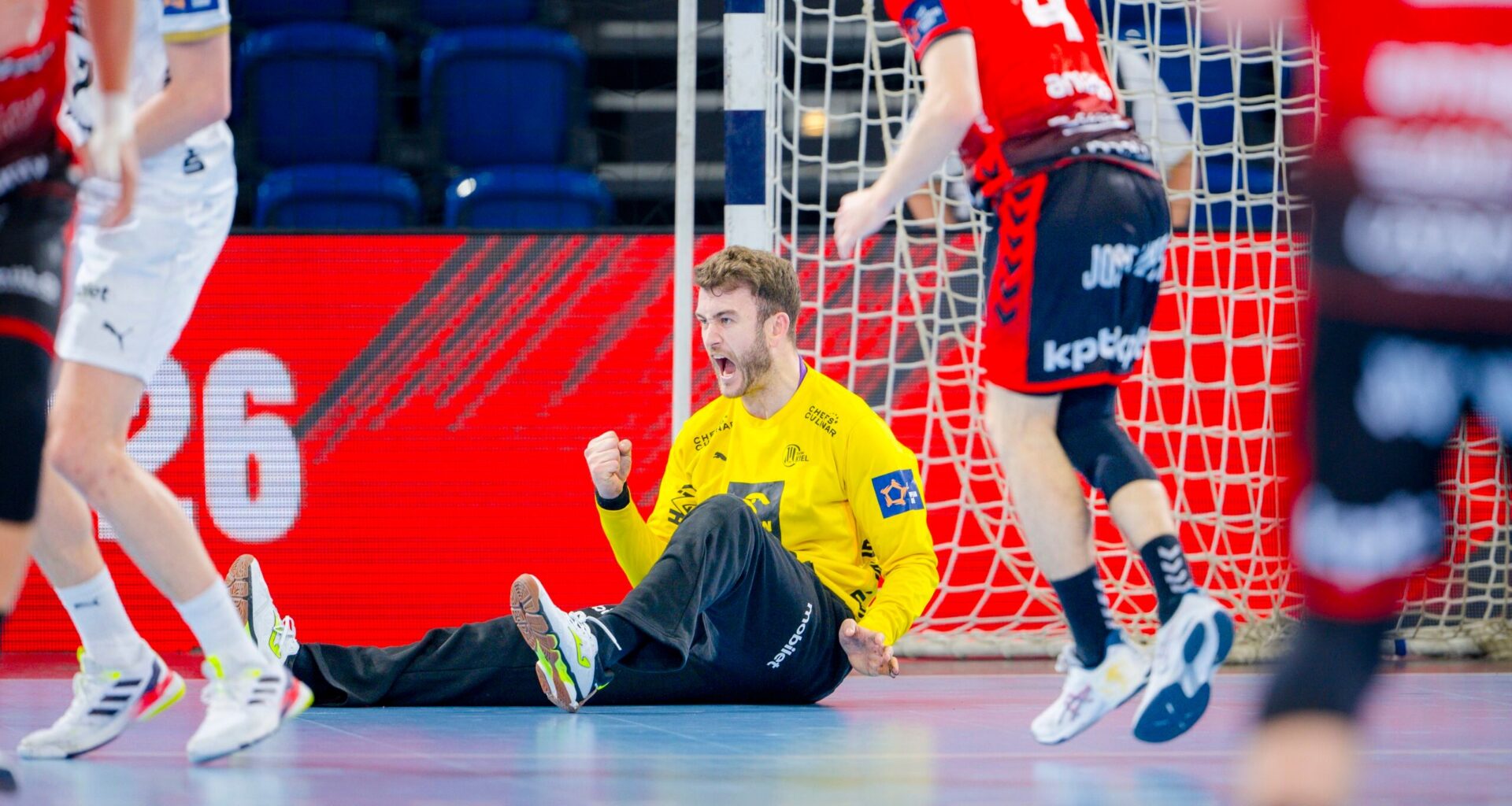 Ein Handballtorwart im gelben Trikot sitzt mit geballter Faust auf dem Boden und feiert, während Spieler in roten und weißen Trikots während des THW Kiel Handball-Derbys in der EHF European League Saison 2025/2026 in der Nähe laufen.