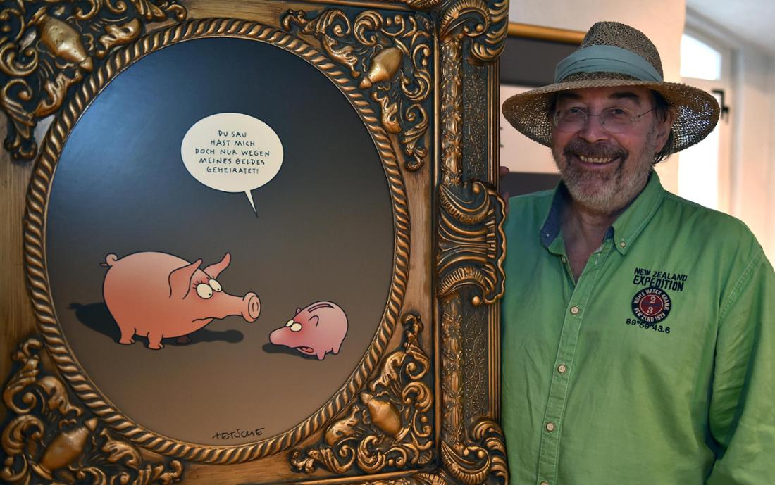 Fred Tödter, bekannt als Cartoonist „Tetsche“, trägt ein grünes Hemd und einen Strohhut und steht neben eines seiner lustigen Bilder-Cartoons im Ausstellungsraum der Burg zu Hagen in Niedersachsen.