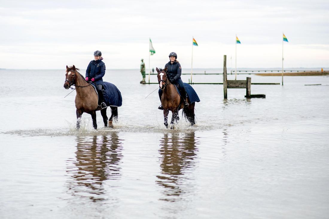 Zwei Frauen reiten auf ihren Pferden im flachen Wasser der Nordsee am Strand entlang. Im Hintergrund ist auf dem Holzsteg am Jadebusen die Skulptur „Jade 2“ des Künstlers Anatol Herzfeld schemenhaft zu sehen.