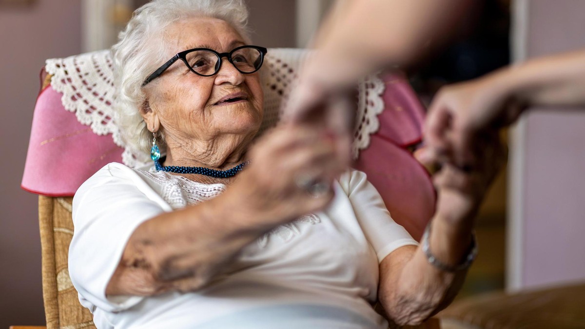 Bei durchschnittlich 5023 Euro im Monat liegen aktuell die Gesamtkosten für einen Platz in einem Berliner Pflegeheim, berechnet die AOK. Der Eigenanteil liege derzeit bei 2.427 Euro. Female home carer supporting old woman to stand up from the armchair at care home