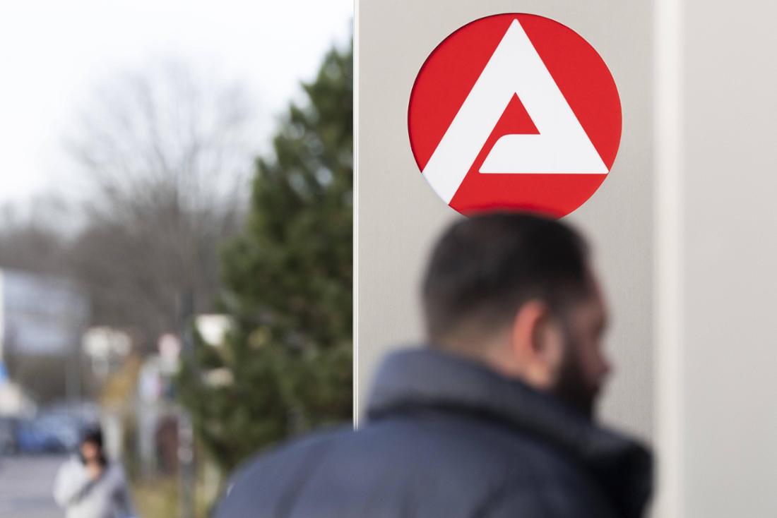 Stele mit dem Logo der Agentur für Arbeit vor dem Gebäude eines Jobcenters. Im Vordergrund sind Umrisse eines Mannes zu sehen.