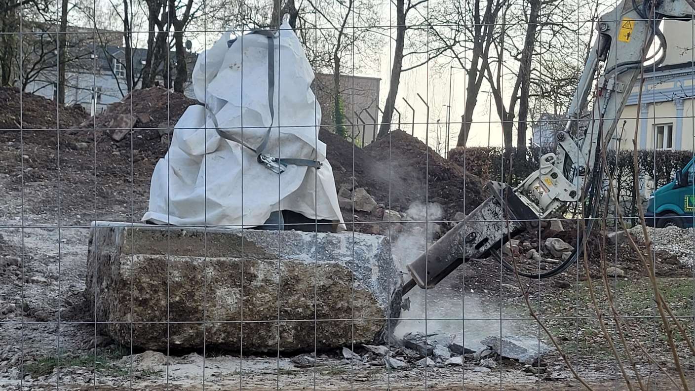 Die neun Zentner schwere Plastik „Der Zeitungsleser“ wird von einem eigens gegossenen Betonfundament getragen. Um einen Transport zu ermöglichen, wird Beton mühsam abgemeißelt.