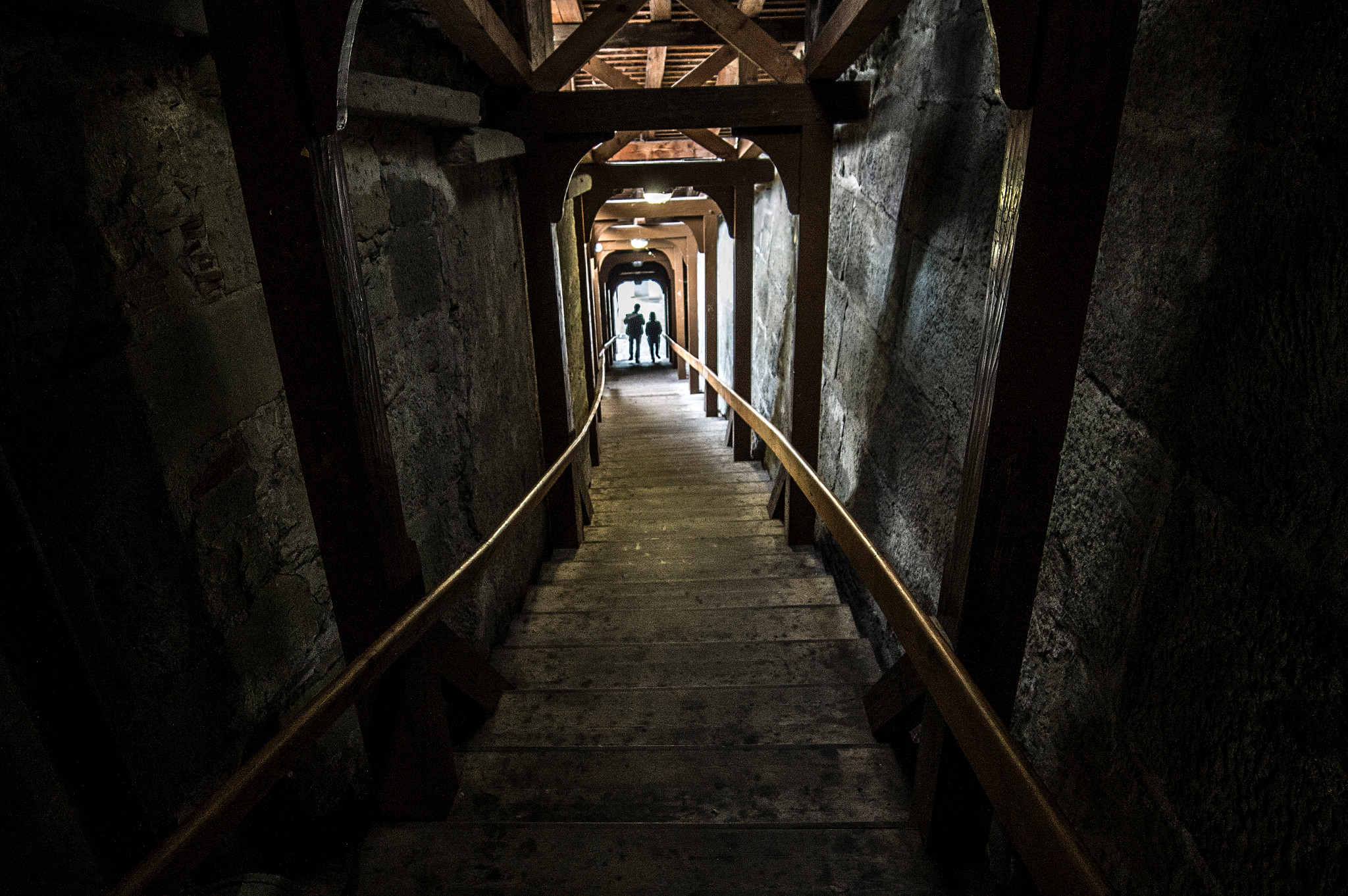 Eine dunkle Holztreppe namens Fricktreppe mit Geländern, die steil nach unten führt, umgeben von Steinwänden mit Personen im Hintergrund.