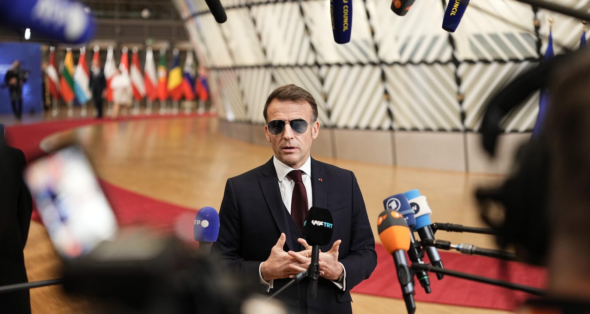 Der französische Präsident Emmanuel Macron trägt im Vorfeld eines Sondergipfels des Europäischen Rates eine Sonnenbrille und spricht in zahlreiche Mikrofone. Im Hintergrund bunte Flaggen.