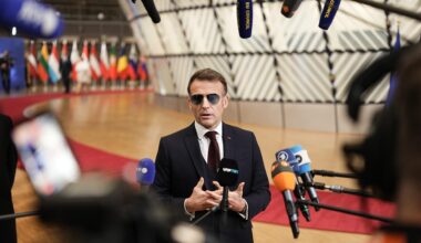 Der französische Präsident Emmanuel Macron trägt im Vorfeld eines Sondergipfels des Europäischen Rates eine Sonnenbrille und spricht in zahlreiche Mikrofone. Im Hintergrund bunte Flaggen.