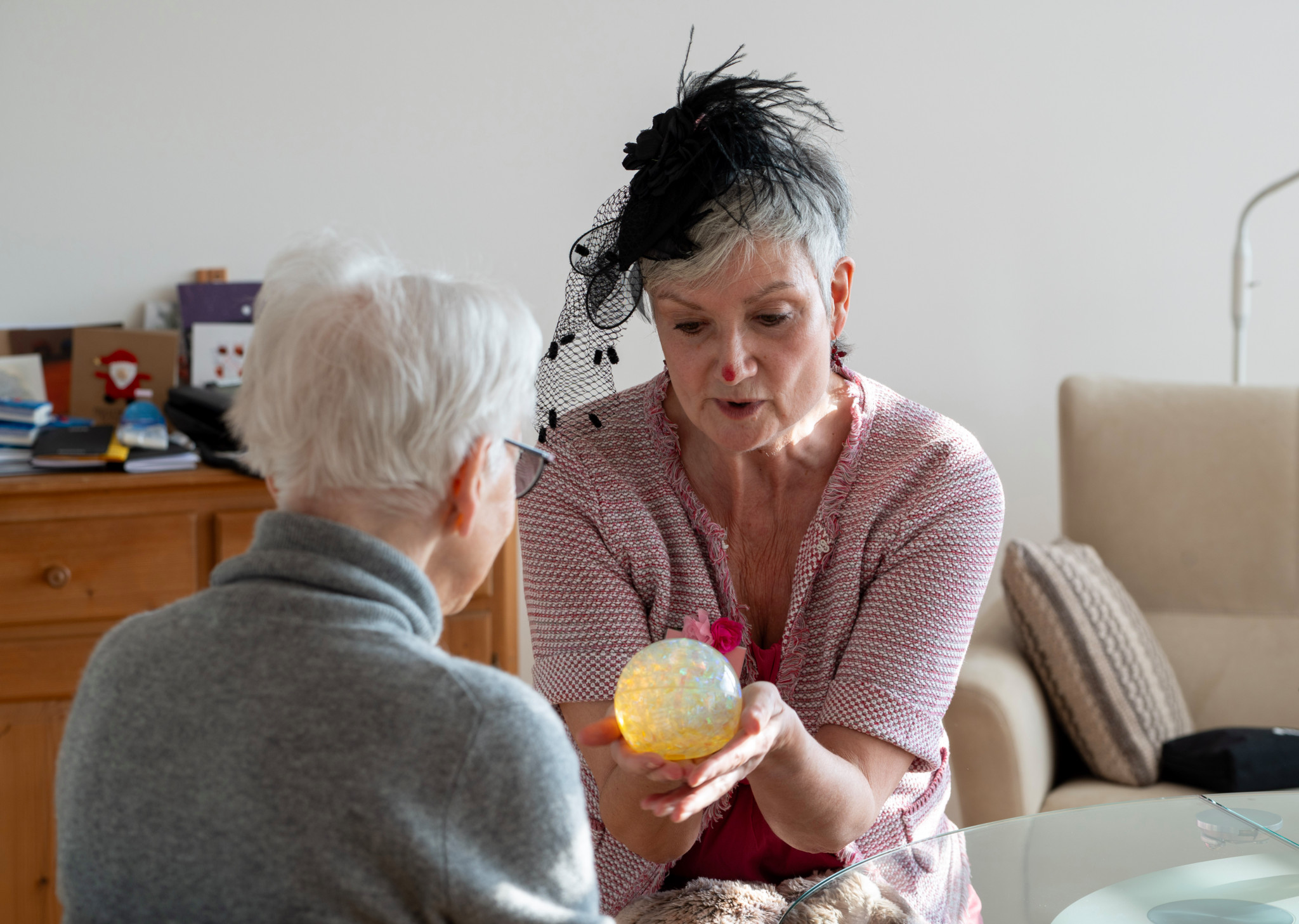 Zwei ältere Frauen unterhalten sich in einem Wohnzimmer, eine von ihnen hält eine glitzernde Glaskugel.