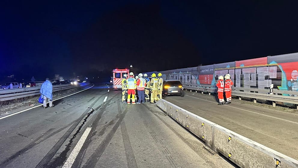 Auf dem Asphalt ist das Glatteis deutlich zu erkennen, ein unverletzter Unfallbeteiligter (links) schützt sich mit einer Decke vor der Kälte