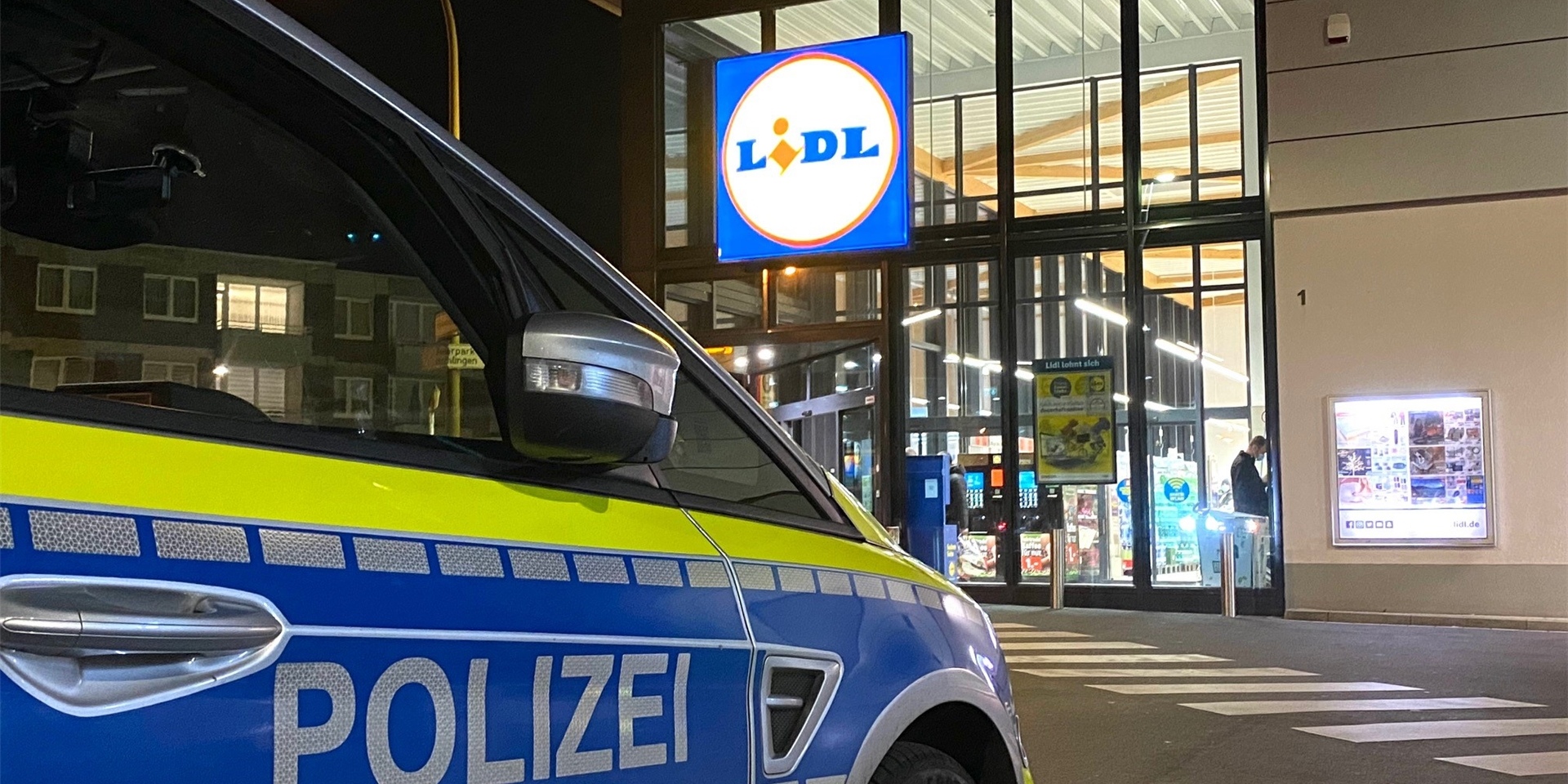 Polizeifahrzeug steht vor einem Lidl-Markt, der nachts beleuchtet ist, während an der Eingangstür Personen vorbeigehen.