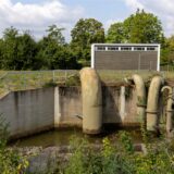 Auf dem Gelände eines Wasserpumpwerks in Recklinghausen sind große, nach unten gebogene Rohre zu sehen, die über einem kleinen Gewässer hängen.