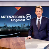 Aktenzeichen XY-Moderator Rudi Cerne steht im Studio und lächelt in die Kamera.
