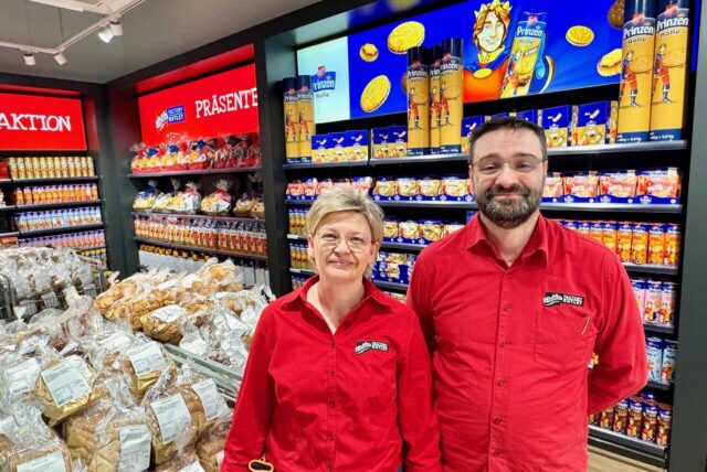 Melanie Homann, die stellvertretende Teamleiterin des Outlet, und der Teamleiter Emilio Lopez in roten Hemden stehen in einem Keks-Outlet in Aplerbeck vor voll bestückten Regalen mit Gebäckprodukten.