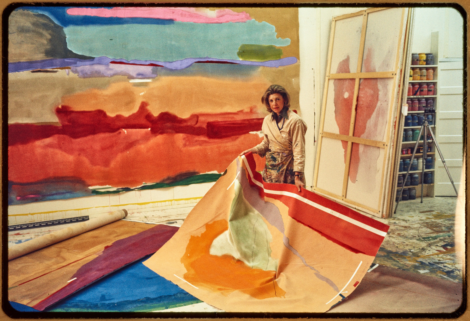 Helen Frankenthaler hält ein abstraktes Gemälde in einem Atelier voller grossformatiger Malereien und Farben.