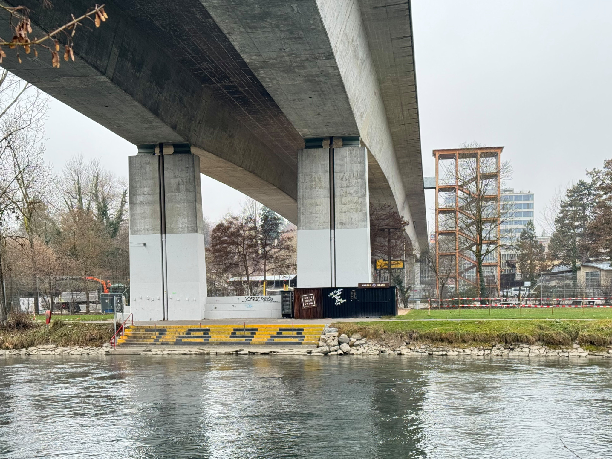 Brücke über einen Fluss mit grauem Betonpfeiler und Treppe. Im Hintergrund sind Bäume und Gebäude zu sehen.