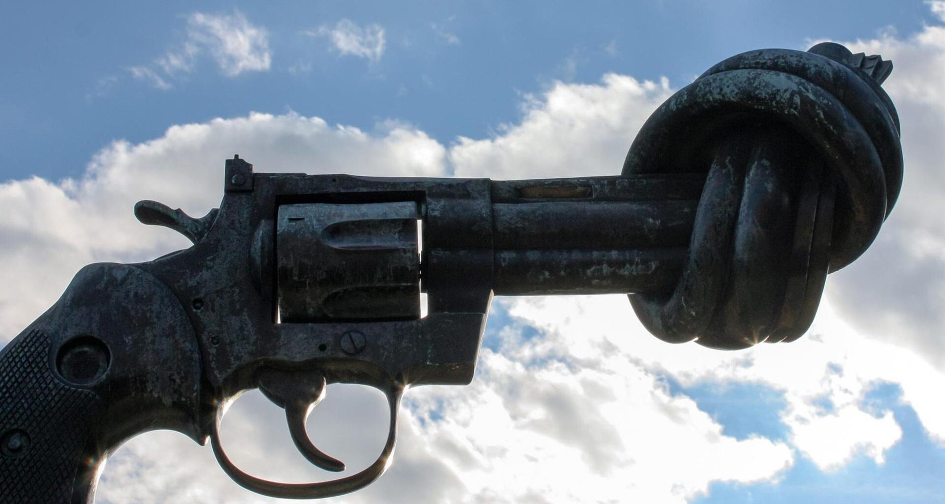 Blick auf die überdimensionale Pistole aus metall mit dem verknoteten Lauf vor einem blauen Himmel mit Wolken.