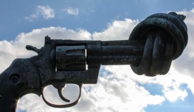 Blick auf die überdimensionale Pistole aus metall mit dem verknoteten Lauf vor einem blauen Himmel mit Wolken.