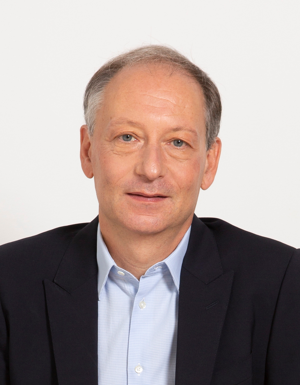 Ein Mann mittleren Alters in einem dunklen Anzug und blauen Hemd mit einem neutralen Gesichtsausdruck vor einem weissen Hintergrund.