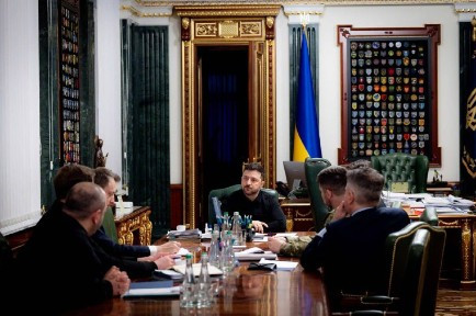 Mann in formellem Büro diskutiert mit mehreren Personen um einen Tisch. Ukrainische Flagge im Hintergrund.