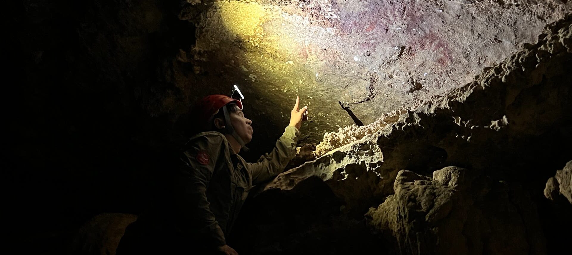 Eine Person mit Helm und Stirnlampe untersucht eine Höhlenwand. Sie zeigt auf eine Stelle, die von der Lampe beleuchtet wird. Die Umgebung ist dunkel und felsig, mit unregelmäßigen Oberflächen und Schatten. Die Szene vermittelt den Eindruck einer wissenschaftlichen Erkundung oder archäologischen Untersuchung in einer Höhle.