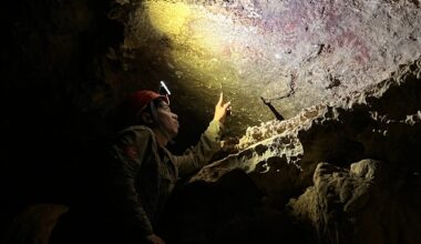 Eine Person mit Helm und Stirnlampe untersucht eine Höhlenwand. Sie zeigt auf eine Stelle, die von der Lampe beleuchtet wird. Die Umgebung ist dunkel und felsig, mit unregelmäßigen Oberflächen und Schatten. Die Szene vermittelt den Eindruck einer wissenschaftlichen Erkundung oder archäologischen Untersuchung in einer Höhle.
