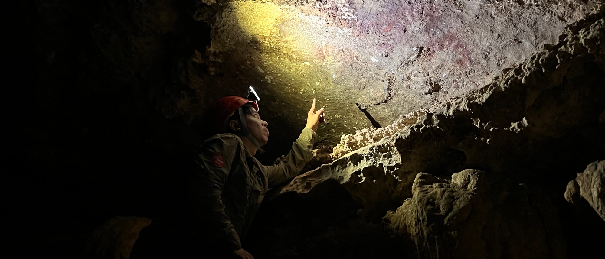 Eine Person mit Helm und Stirnlampe untersucht eine Höhlenwand. Sie zeigt auf eine Stelle, die von der Lampe beleuchtet wird. Die Umgebung ist dunkel und felsig, mit unregelmäßigen Oberflächen und Schatten. Die Szene vermittelt den Eindruck einer wissenschaftlichen Erkundung oder archäologischen Untersuchung in einer Höhle.