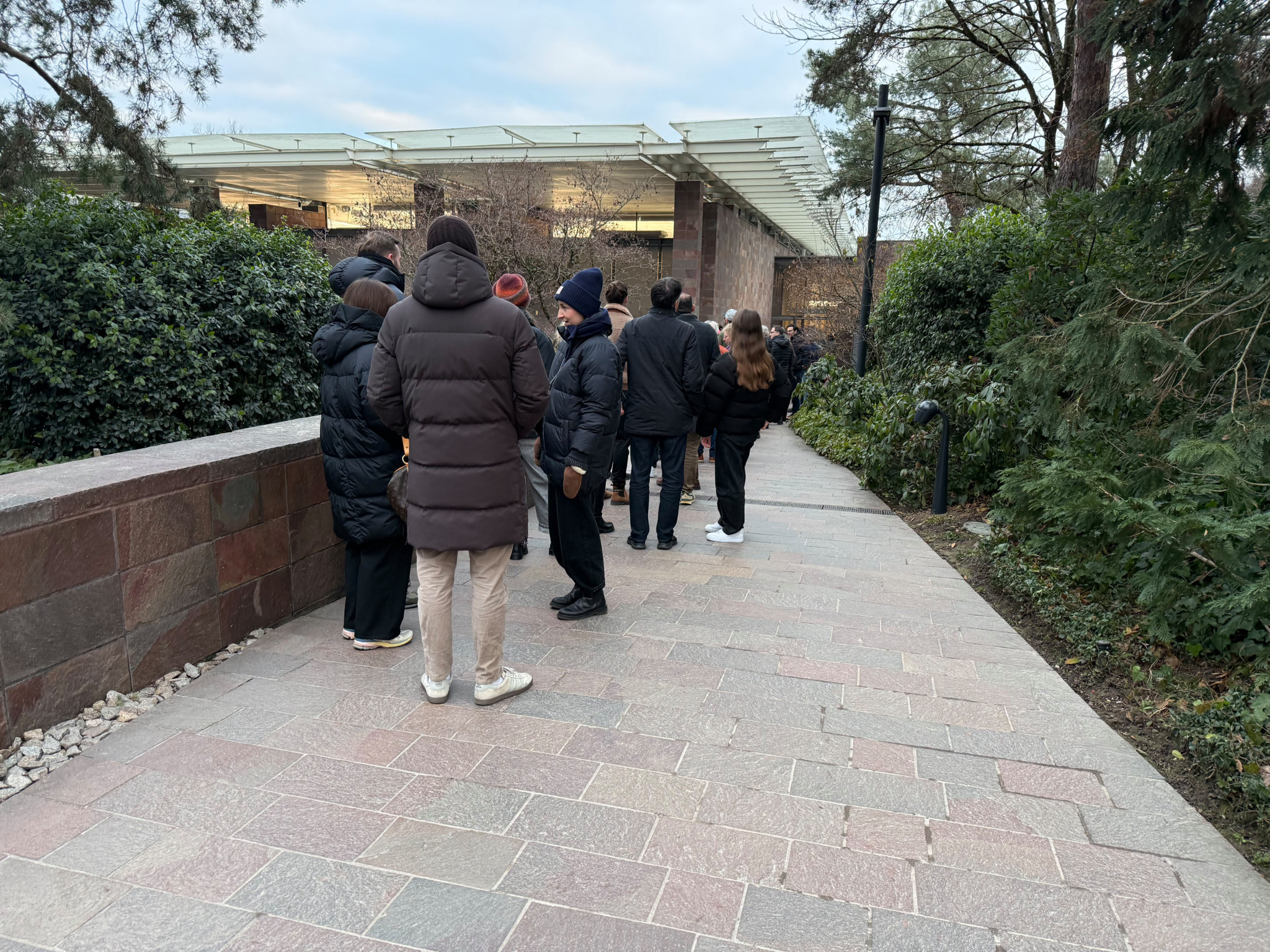 Gruppe von Menschen in Winterkleidung steht auf einem gepflasterten Weg, umgeben von Bäumen und Büschen.