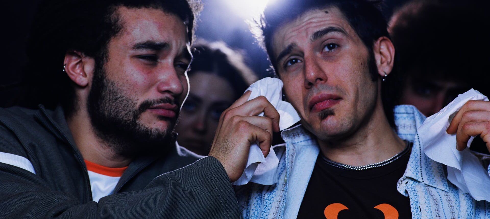Zwei junge Männer sitzen nebeneinander im Kino; einer trocknet dem anderen mit einem Taschentuch Tränen aus dem Gesicht. Beide wirken emotional bewegt.