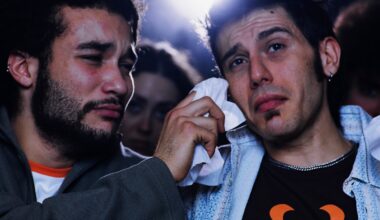 Zwei junge Männer sitzen nebeneinander im Kino; einer trocknet dem anderen mit einem Taschentuch Tränen aus dem Gesicht. Beide wirken emotional bewegt.