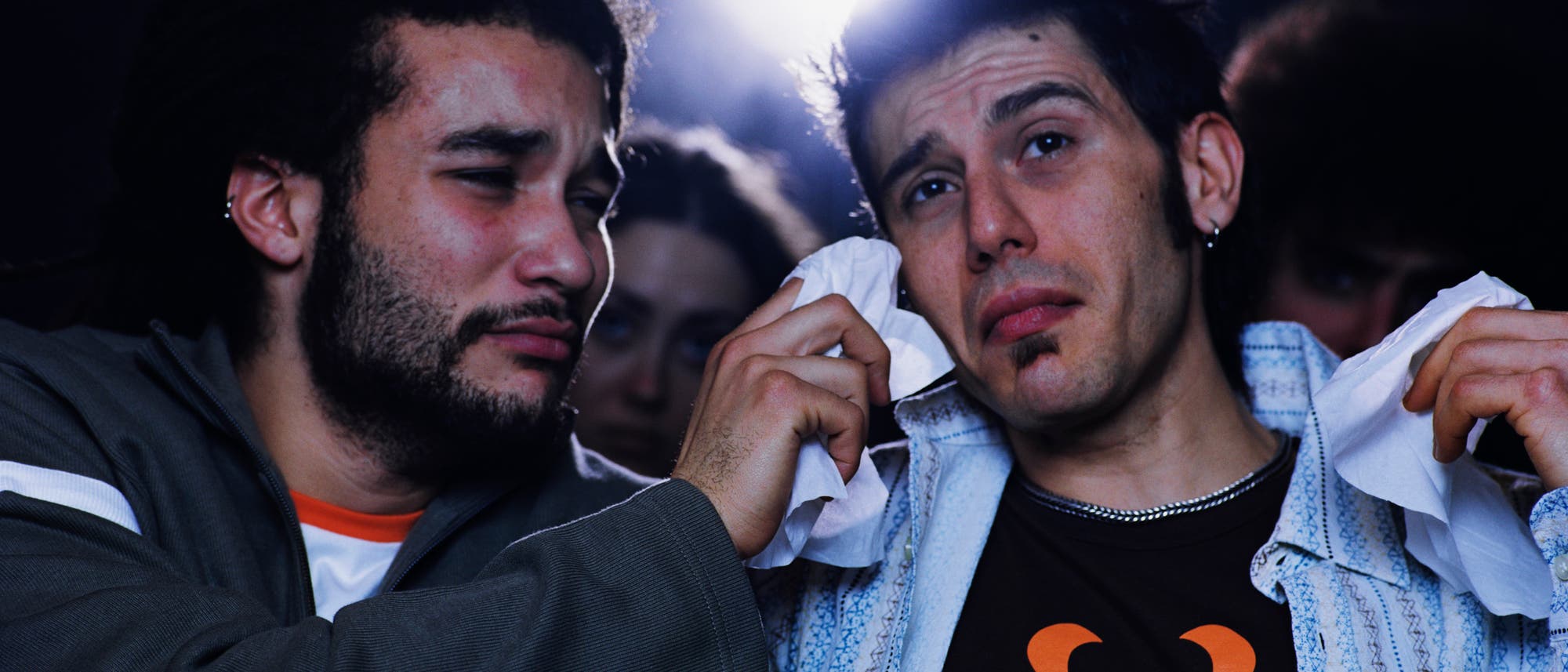 Zwei junge Männer sitzen nebeneinander im Kino; einer trocknet dem anderen mit einem Taschentuch Tränen aus dem Gesicht. Beide wirken emotional bewegt.