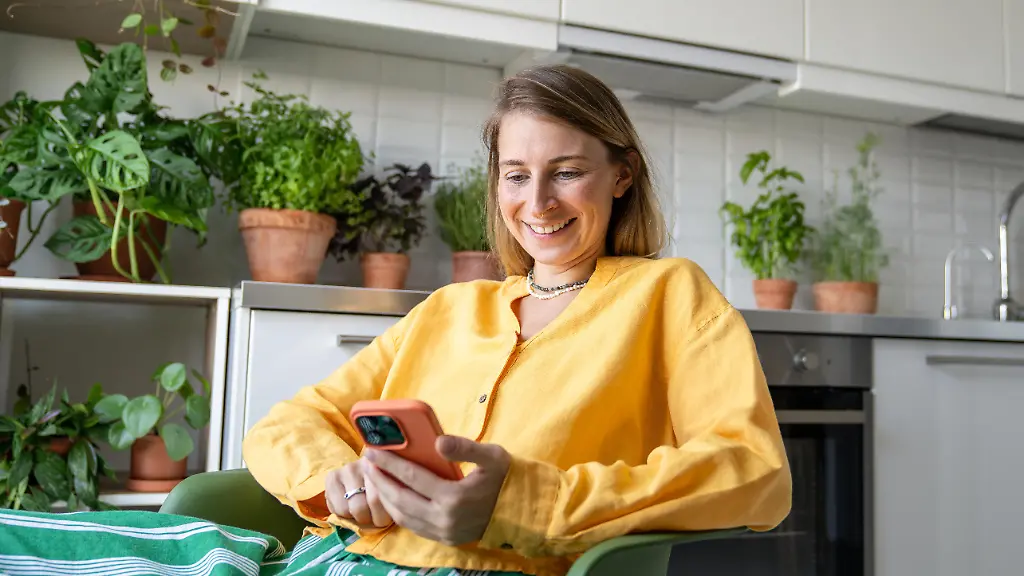 Laughing-woman-engrossed-in-smartphone-watching-video-news-and-social-feed-sit-in-kitchen-surrounded-by-plants
