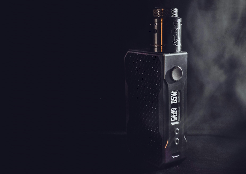 Ein schwarzer E-Zigaretten Akkuträger (Box Mod) mit einem beleuchteten Display, das Einstellungen anzeigt, und einem aufgeschraubten Tankverdampfer vor dunklem Hintergrund mit leichtem Dampfnebel