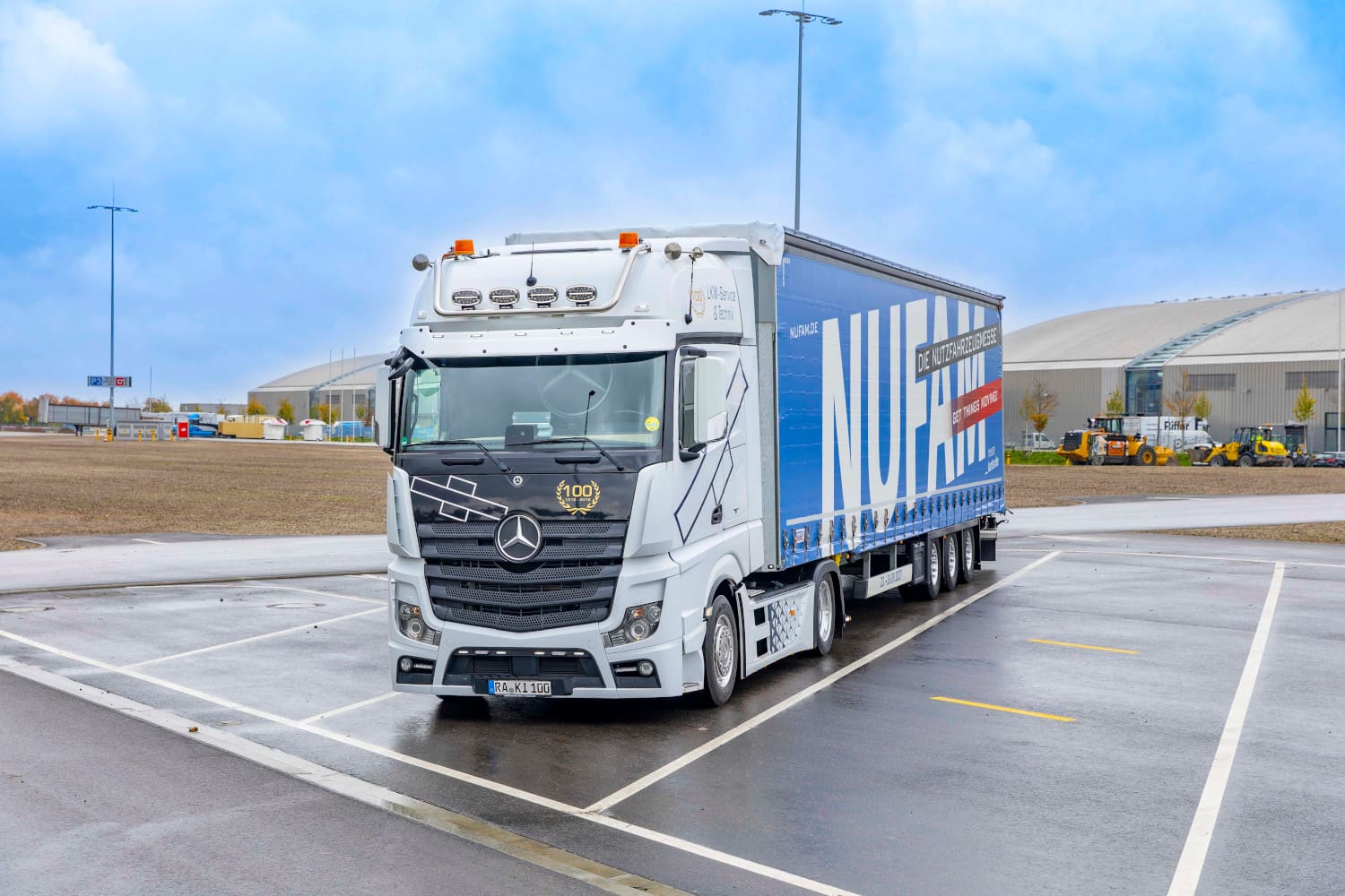 Ein wei&szlig;er Mercedes-Benz Sattelzug steht auf dem Au&szlig;engel&auml;nde der Messe Karlsruhe. Der blaue Auflieger tr&auml;gt die gro&szlig;fl&auml;chige Aufschrift &bdquo;NUFAM &ndash; Die Nutzfahrzeugmesse&ldquo;. Im Hintergrund sind Messehallen unter einem leicht bew&ouml;lkten Himmel zu sehen.