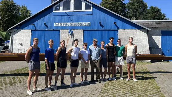 In Ratzeburg wird der Film "Adams Acht" mit Schauspieler Oliver Masucci (5.v.r.) gedreht. | Sören Gerhardt / NDR Neun Männer stehen nebeneinander vor einem blauen Gebäude auf dem steht: Ratzeburger Ruderclub.