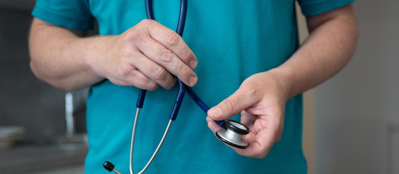 Ein Hausarzt steht in seiner Hausarztpraxis und hält ein Stethoskop.