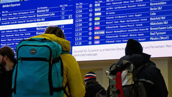 Reisende stehen vor einer Anzeigetafel an einem Bahnhof.