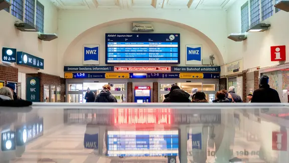 Reisende stehen vor einer Anzeigetafel der Bahn